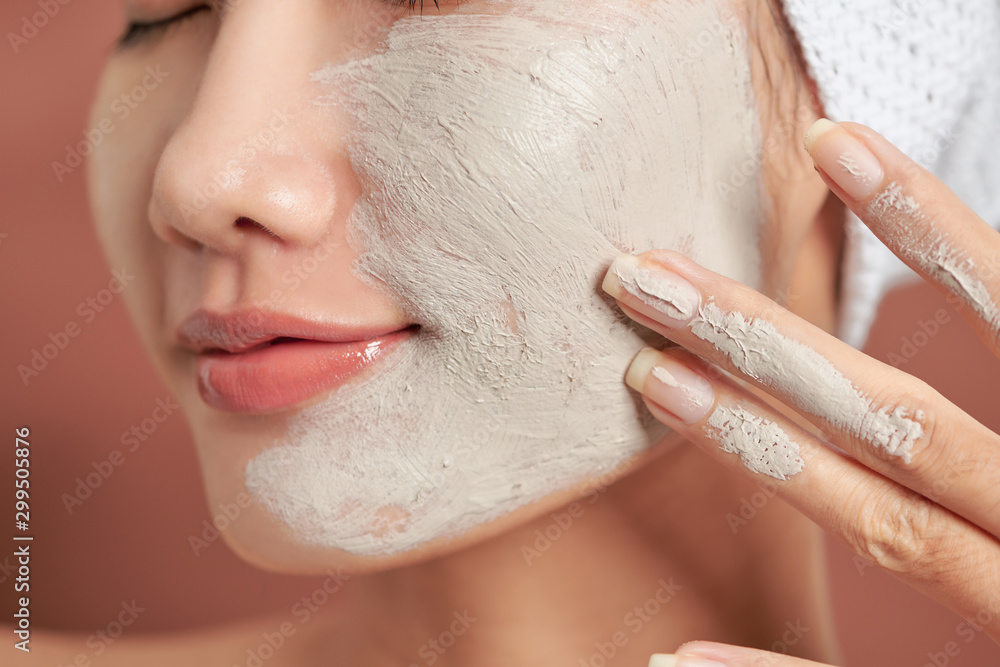 Close-up portrait of attractive girl with a towel on head and clay mask on face isolated over orange