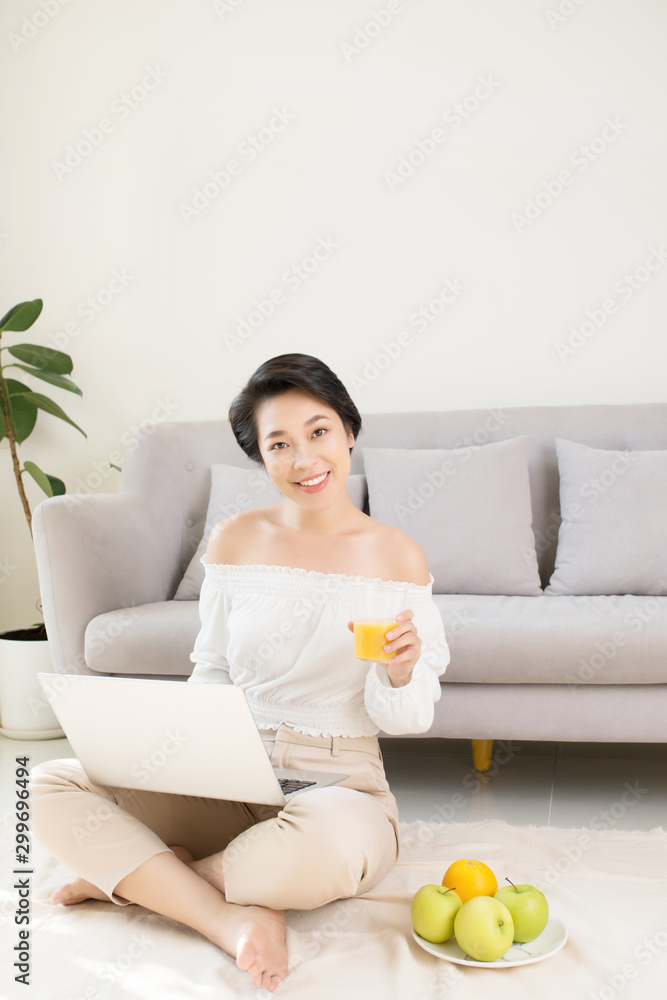 Beautiful Asian woman using laptop while sitting on the floor and drinking juice