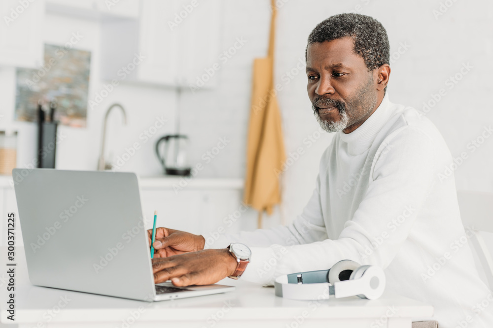 serious african american man using laptop while sitting at table near headphones