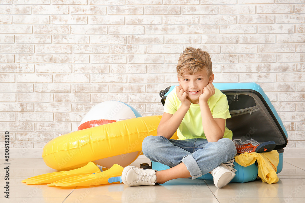 Cute little boy and suitcase with things for summer vacation near brick wall