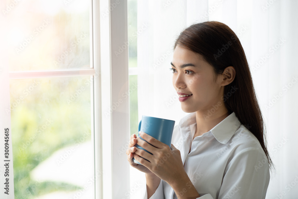 亚洲女人早上醒来后喝着一杯咖啡，透过玻璃窗看