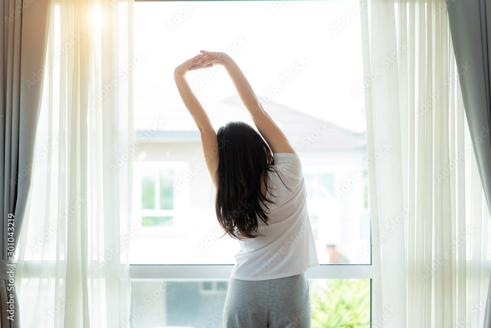 Rear view of Asian Woman stretching hands and body near window after wake up in bedroom at home. Con