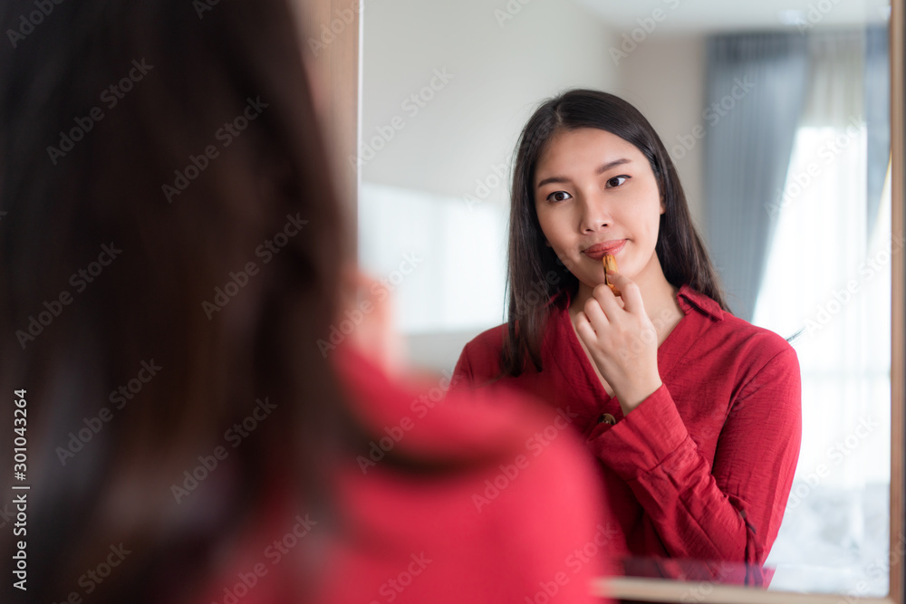 美丽的亚洲女人穿着红色衣服，在家里的卧室里对着镜子涂口红。