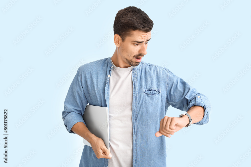 Stressed man with laptop looking at his watch on light background