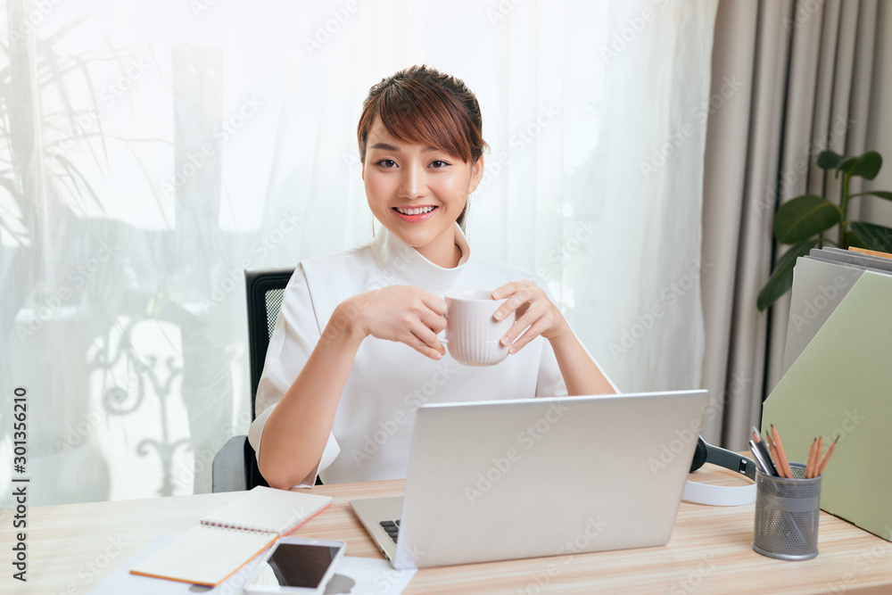 漂亮的年轻亚洲女人在家用笔记本电脑工作时喝茶或喝咖啡