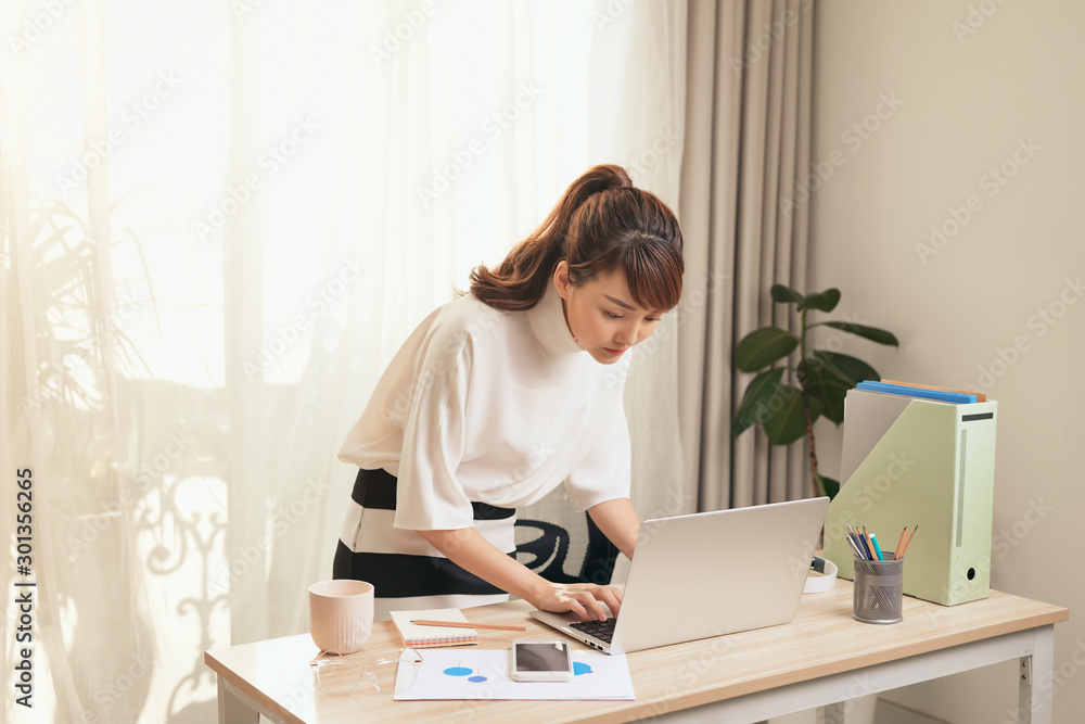 Young Asian businesswoman working with laptop and standing behind the desk at home.