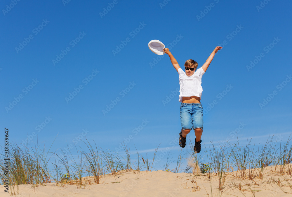 Happy boy jump high on the sand dune holding hat