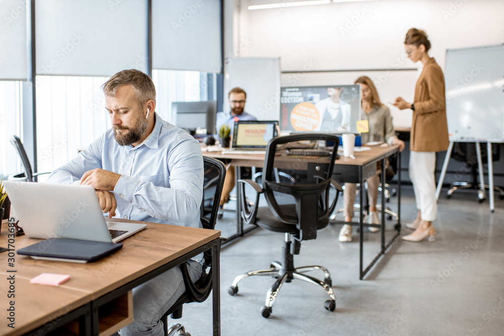 Senior man dressed casually working on the laptop in the office with coworkers on the background