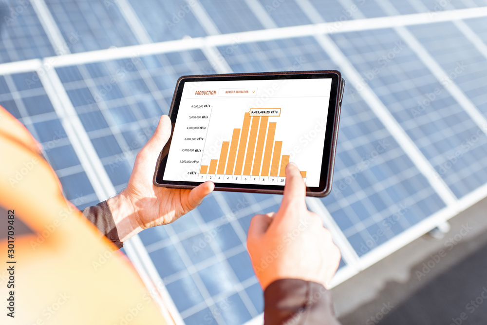 Workman examining genaration of solar power plant, holding digital tablet with a chart of electricit