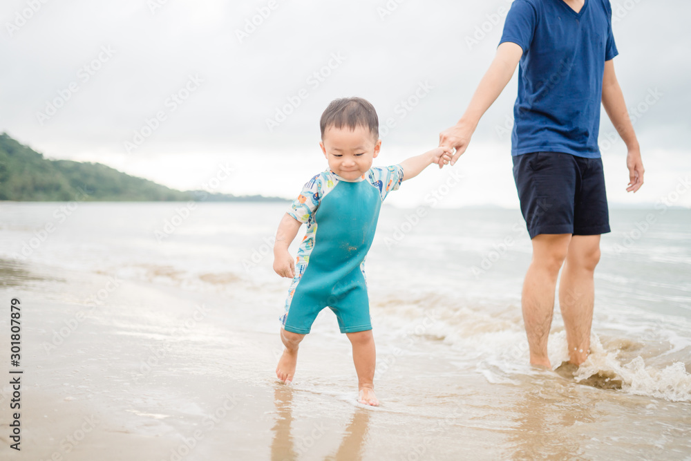 亚洲一个蹒跚学步的男孩和他的父亲一起走向大海。父亲牵着他的儿子游泳和玩耍。