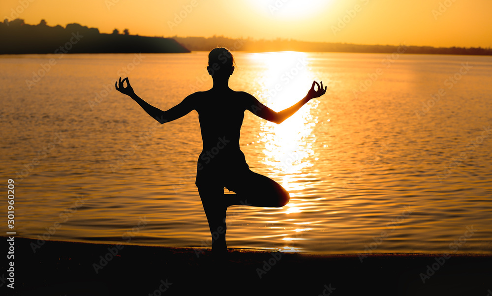 Beautiful young woman practicing yoga near river at sunset