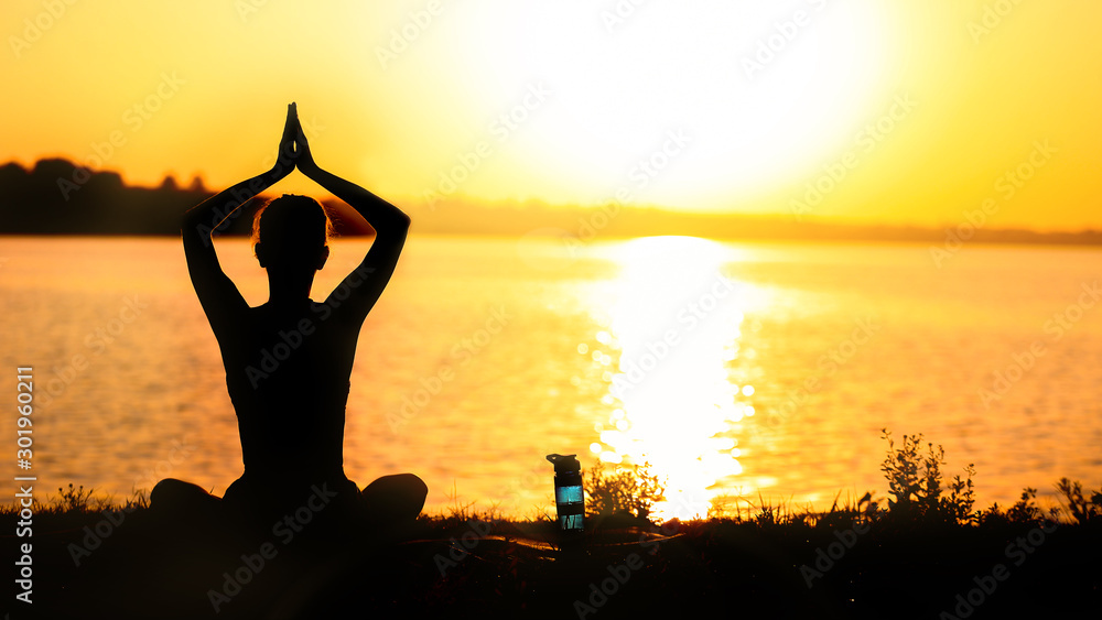 Beautiful young woman practicing yoga near river at sunset