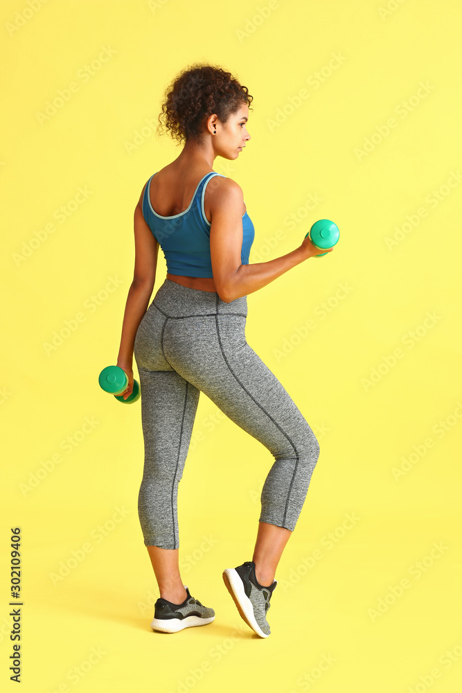 Sporty African-American woman with dumbbells on color background