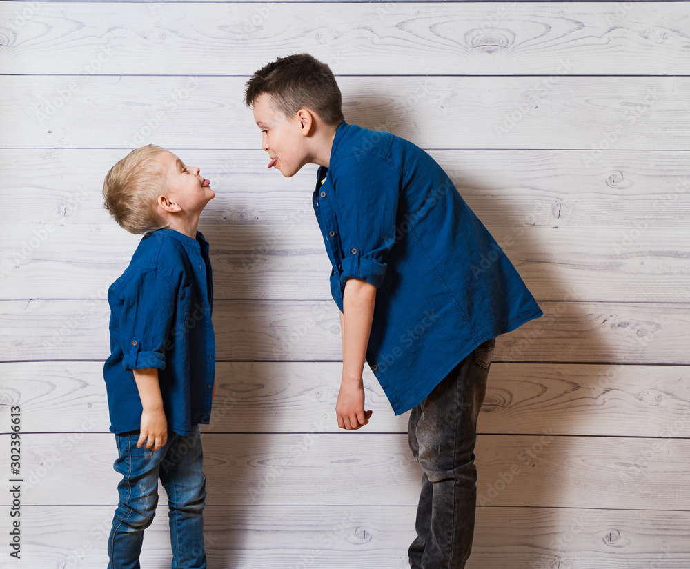 Two boys in casual clothes in white wooden background showing tongues to each other. Two brothers pl