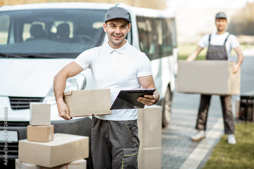 Portrait of delivery man in uniform standing with documents near the cargo van vehicle, colleague un