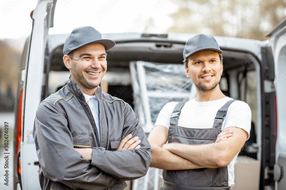 Portrait of a two cheerful delivery men or movers in workwear standing near a cargo vehicle trunk fu