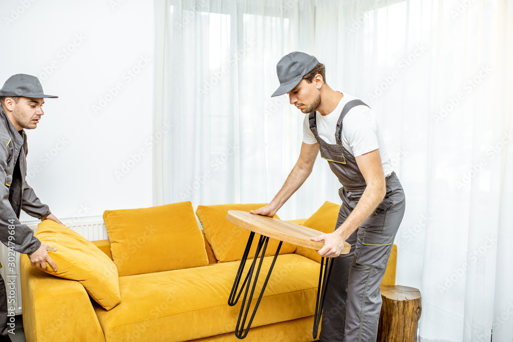 Professional movers in uniform placing furniture at the living room of a new apartment during a relo