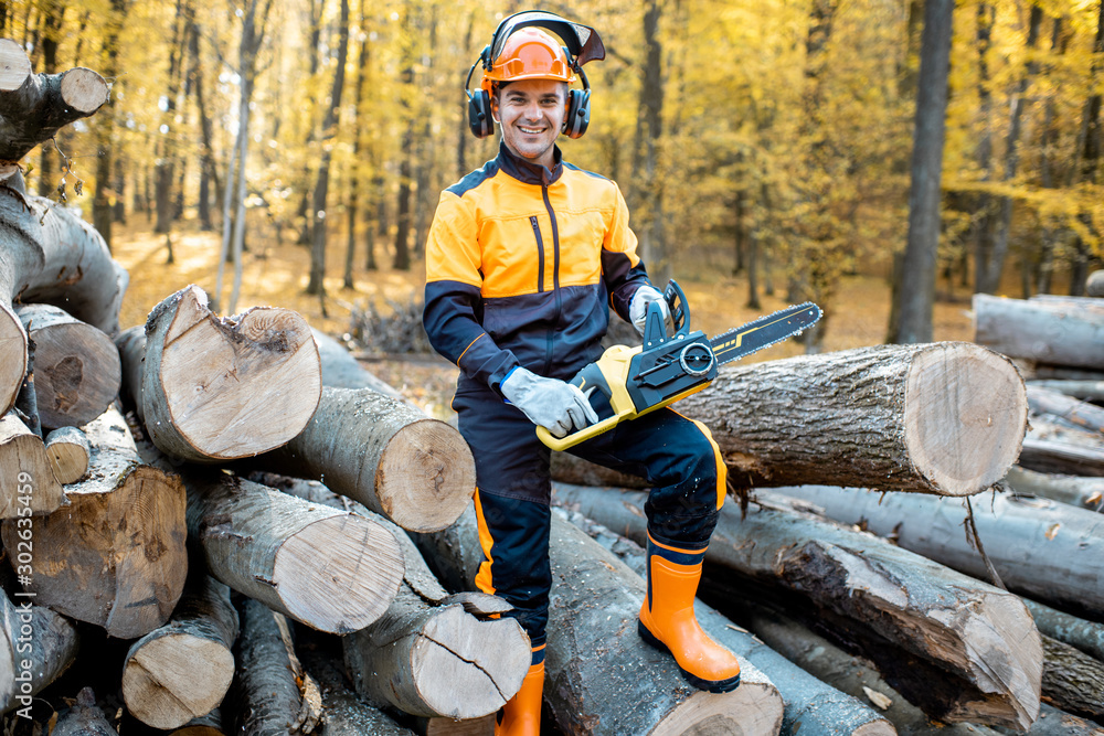 Portrait of a cheerful professional lumberjack in protective workwear standing with a chainsaw on a 