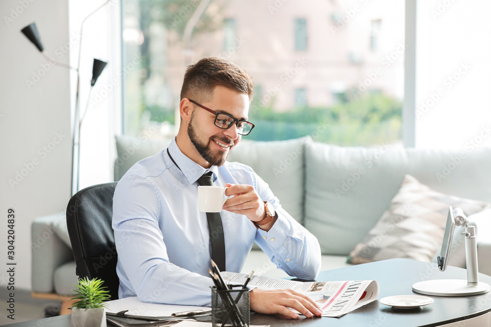 Handsome businessman drinking coffee while reading newspaper in office