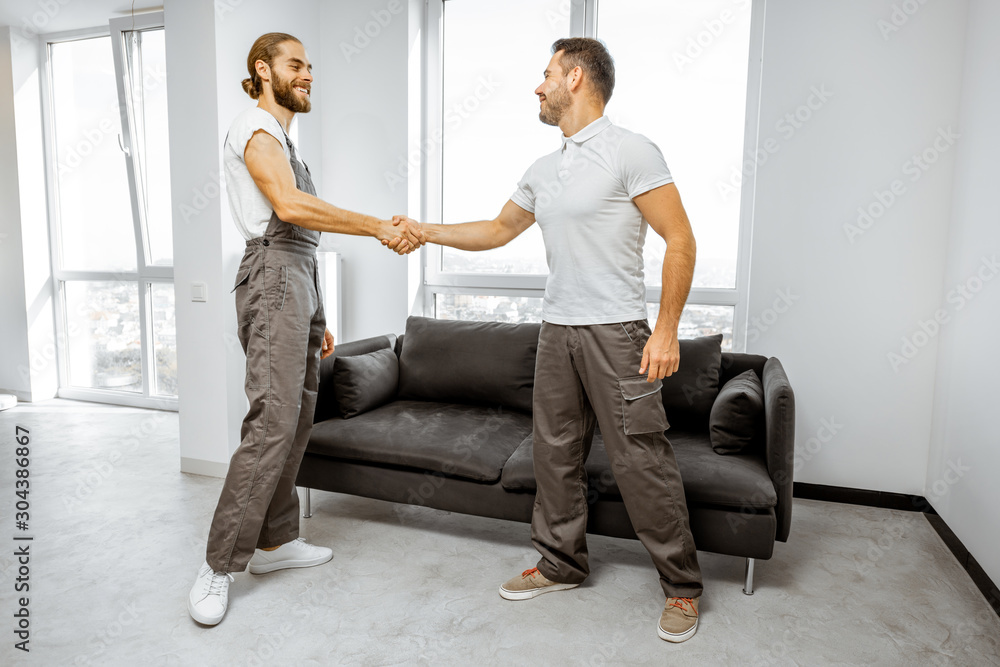 Two happy professional movers or repairmen in workwear shaking hands together, finishing work in the