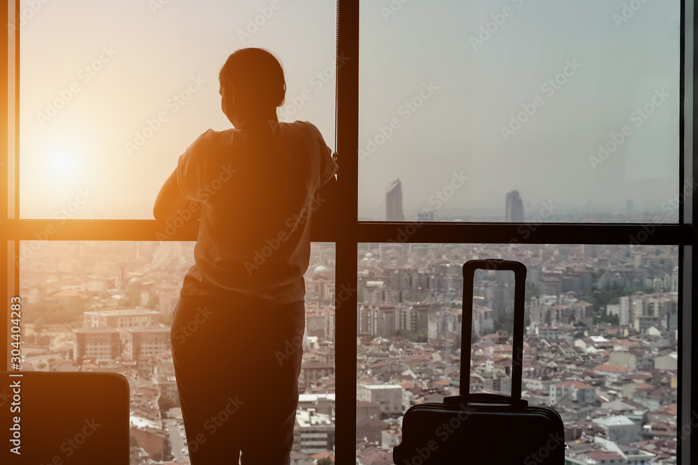 silhouette of young girl traveler with suitcase in hotel room with panoramic city view. She is stand