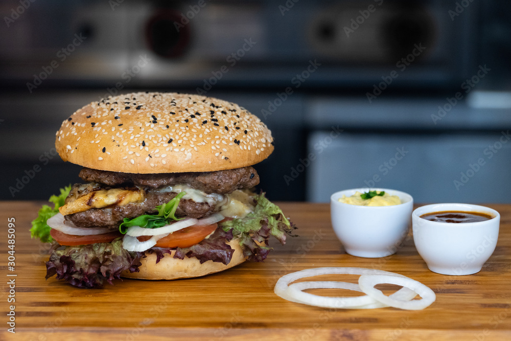 A big fresh colorful burger with meat, egg, salad, onion rings and dips on a wooden tray