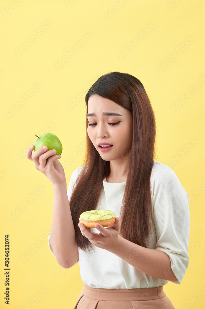 Portrait of a smiling young asian woman choosing between donut and green apple isolated over yellow 