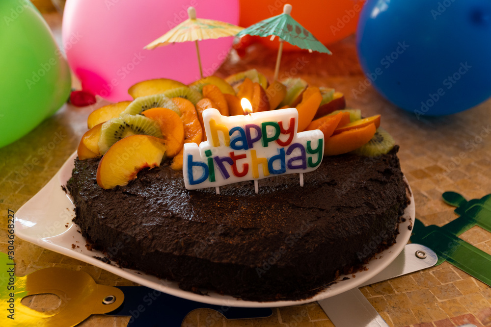 A home made vegan chocolate cake with fresh fruit, small umbrellas, balloons and a candle saying Hap