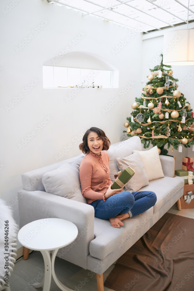 Beautiful asian woman in casual sweater holding gift near decorated christmas tree, greeting card co