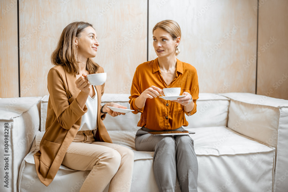 Two female colleagues having cheerful conversation, sitting together on the couch during a coffee br