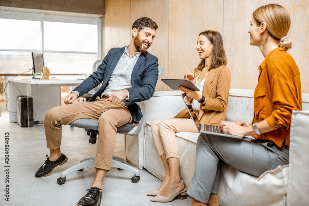 Playful man flirting with a young colleagues, women sitting and working on the couch in the office