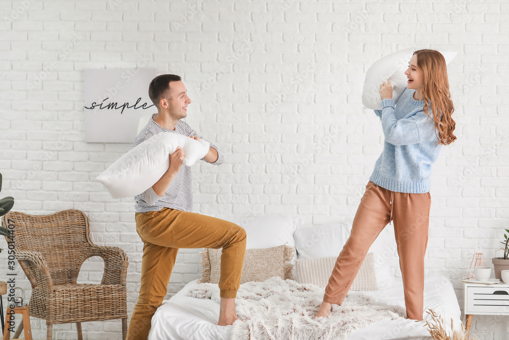 Beautiful young couple fighting with pillows on bed at home