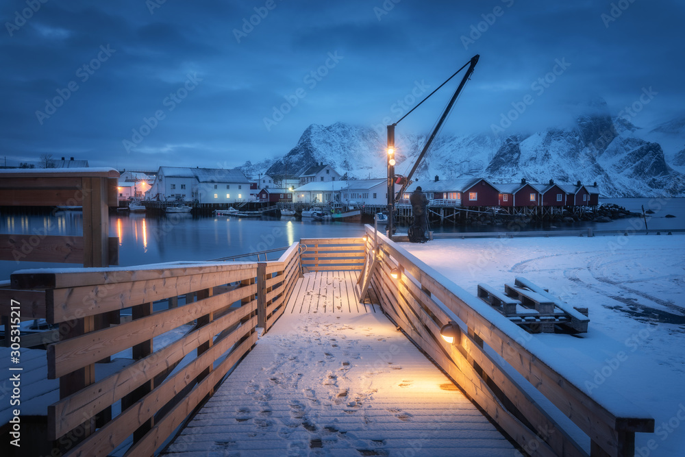 Snowy wooden pier on the sea coast with lights, rorbu and houses, boats and snow covered mountains i