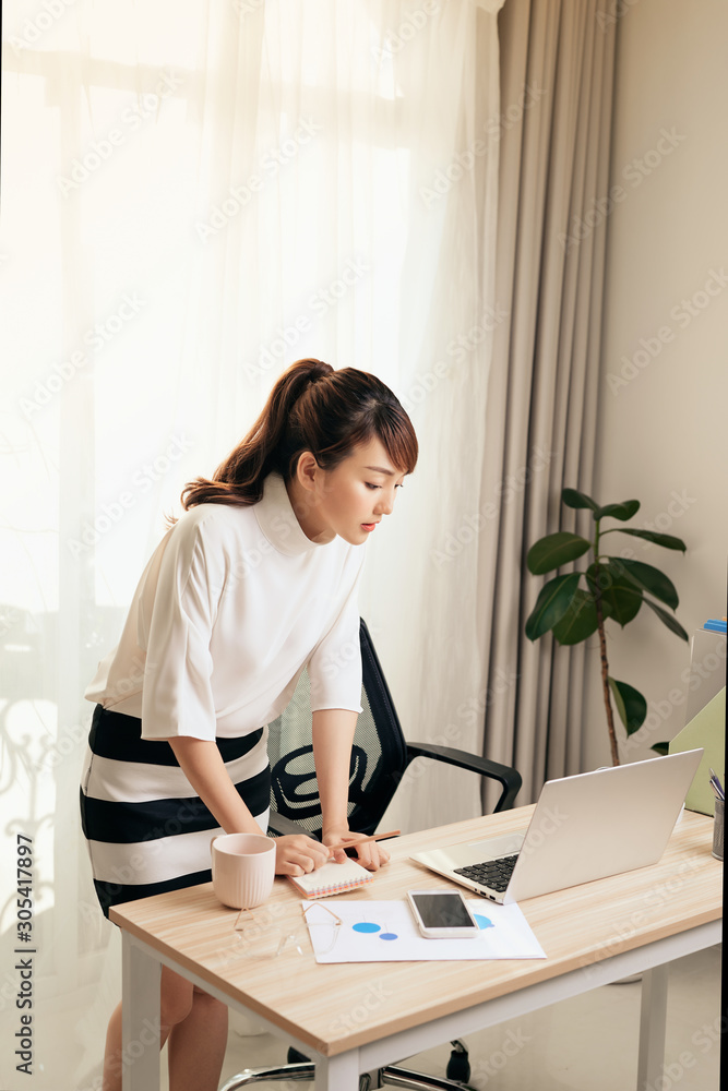 Young Asian businesswoman working with laptop and standing behind the desk at home.