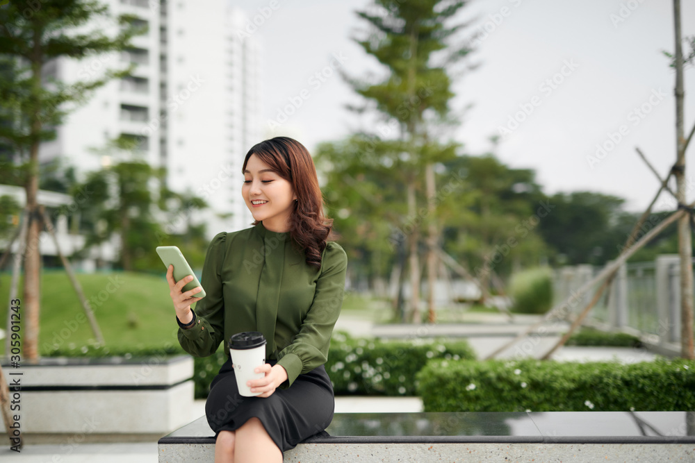 Happy young woman with a disposable coffee cup sitting on the park bench and using her smartphone