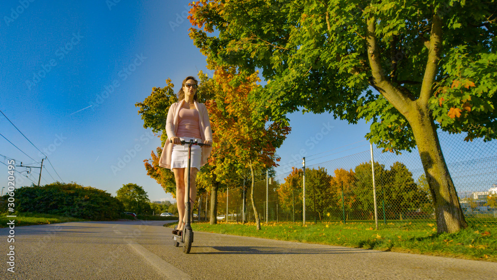 LOW ANGLE: Millennial woman rides an electric scooter down a scenic avenue.