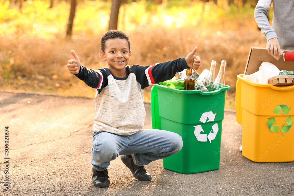 Little African-American boy showing thumb-up near containers for trash outdoors. Concept of recyclin