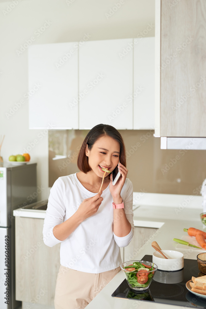 漂亮的年轻女人穿着围裙在家里的厨房里用手机烹饪健康沙拉