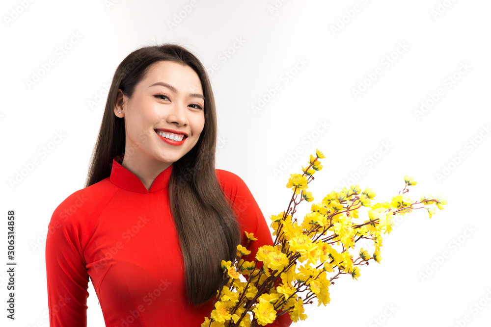 Elegant Asian woman holding peach flowers while wearing Ao dai over white background.