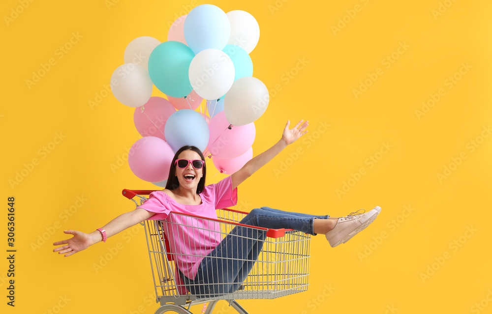 Young woman with shopping cart and balloons on color background