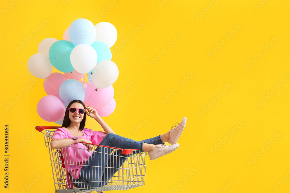 Young woman with shopping cart and balloons on color background