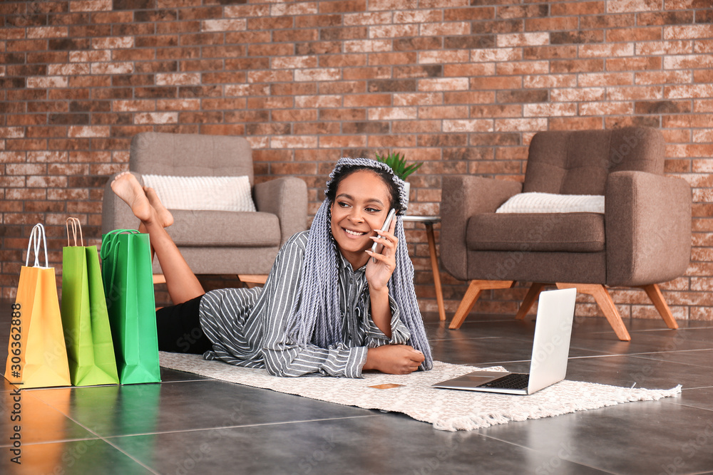 African-American woman talking by phone while using laptop for online shopping at home