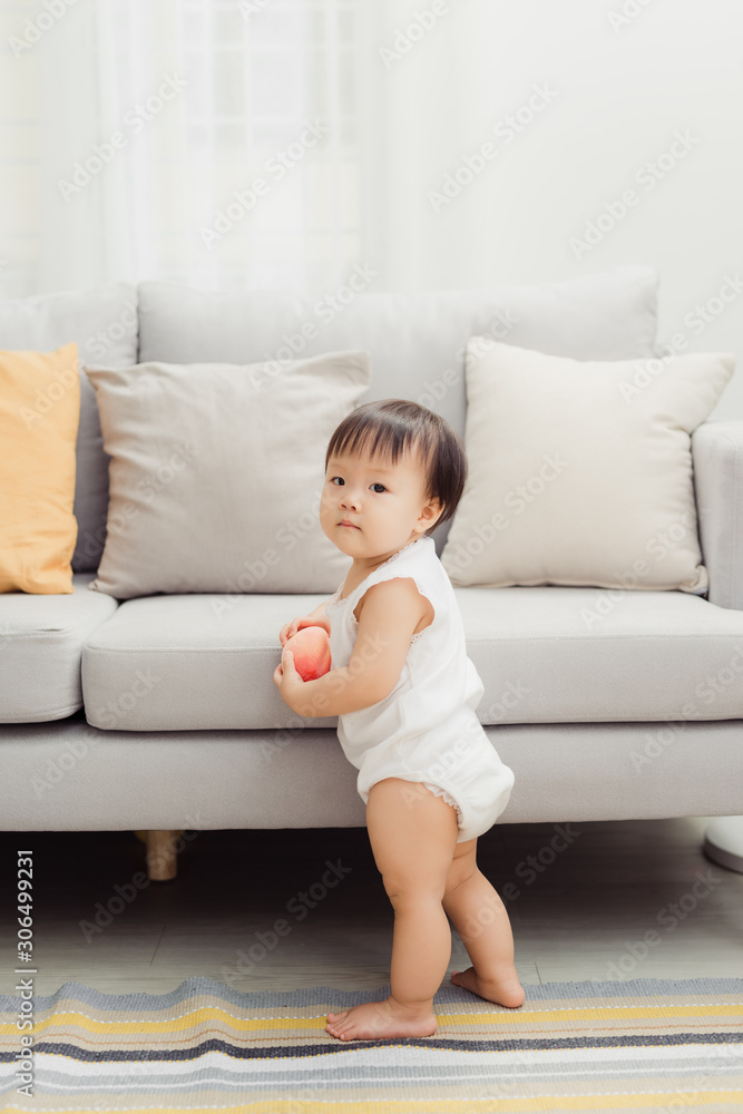 可爱的婴儿站在沙发旁边。可爱的快乐脸女婴画像