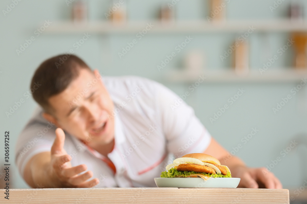 Overweight man stretching hand for burger on kitchen table. Diet concept