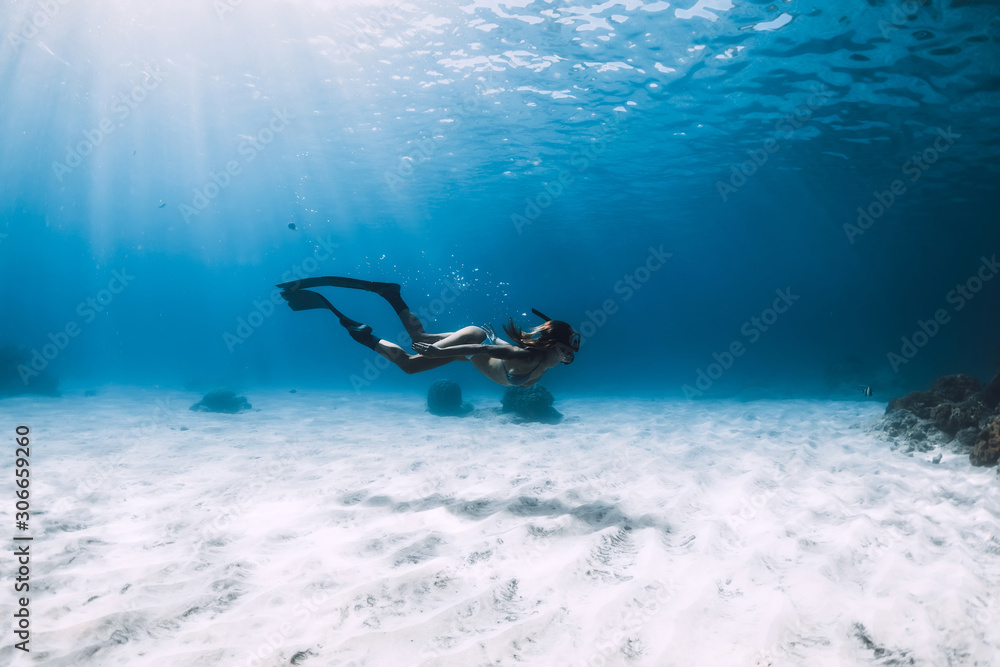 Freediver girl with fins glides over sandy bottom with fishes in blue ocean