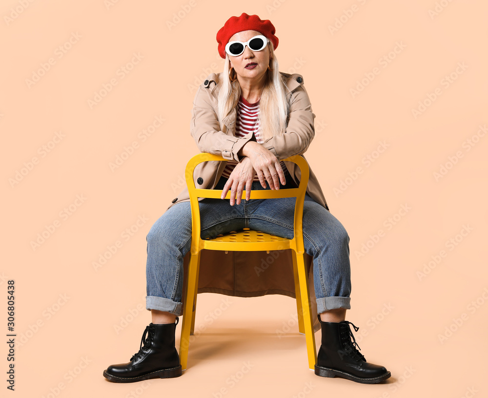 Fashionable mature woman sitting on chair against color background