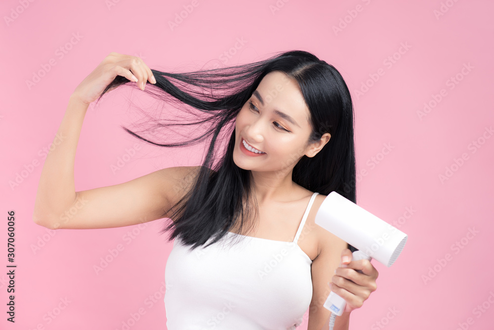 Beautiful Smiling Asian Girl With Black Long Straight Hair Using Hairdryer. Isolated on pink backgro