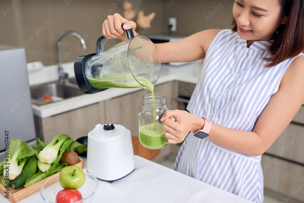 年轻的亚洲女人在现代厨房里把绿色奶昔倒进玻璃里