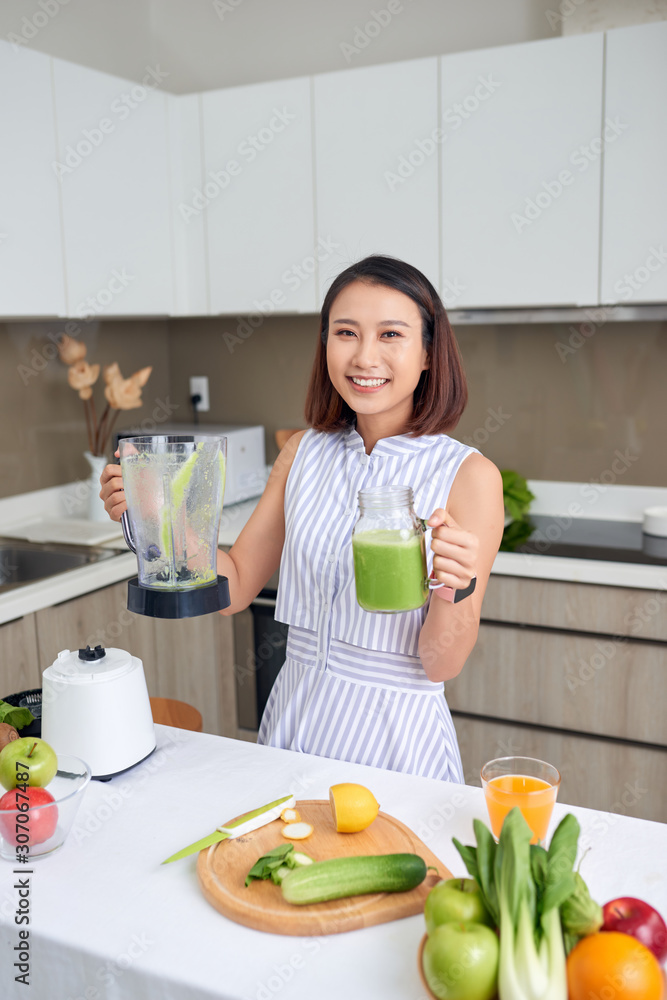 家里厨房里拿着绿色奶昔，面带微笑的年轻亚洲女性。