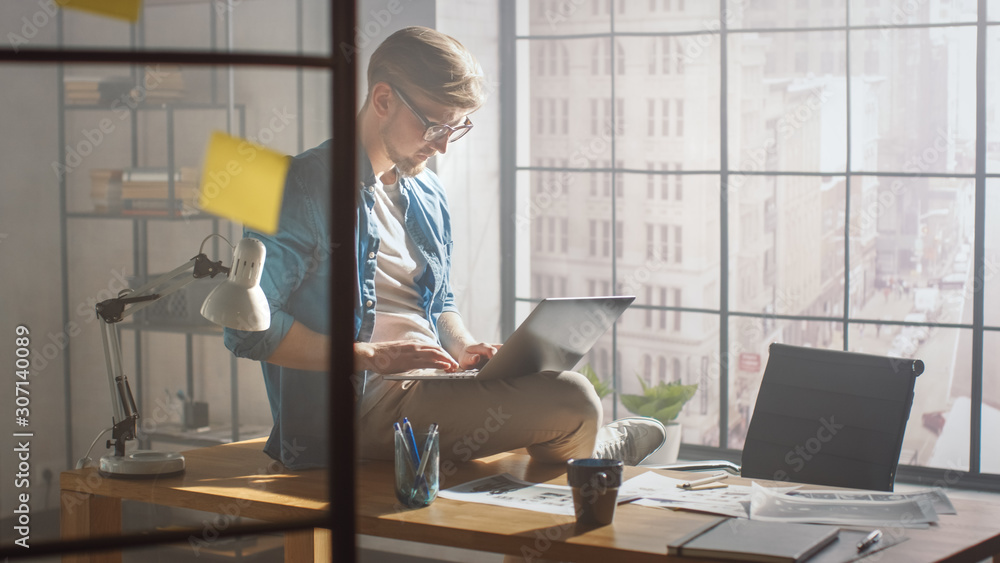 Professional Creative Designer Sits on His Desk Holds Laptop on the Knees and Working on the Project